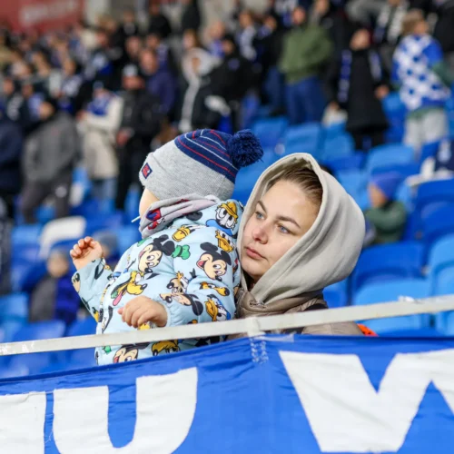Kobieta trzymająca dziecko w różnorodnej odzieży na stadionie, obserwująca wydarzenie sportowe wśród tłumu kibiców.