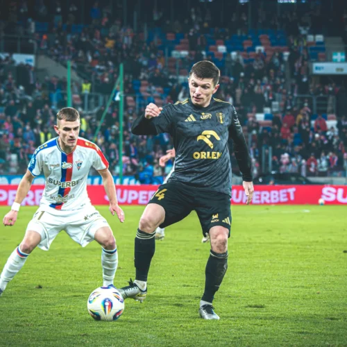 Na zdjęciu przedstawieni są piłkarze podczas meczu, gdzie jeden z zawodników stara się zdobyć piłkę, a w tle widać zapełnione trybuny.