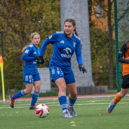 Na zdjęciu widać cztery zawodniczki grające w piłkę nożną, z których dwie są ubrané w niebieskie stroje, a dwie w pomarańczowe, podczas meczu na boisku trawiastym.