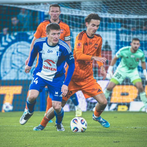 Na zdjęciu widać piłkarzy w trakcie meczu, z jednym zawodnikiem w niebieskim stroju oraz innym w pomarańczowym, chwytających się w rywalizacyjnej akcji na boisku.
