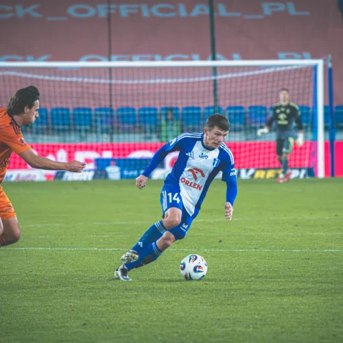 Na zdjęciu dwóch piłkarzy w trakcie dynamicznej akcji na boisku, z jedną osobą starającą się przejąć piłkę od przeciwnika.