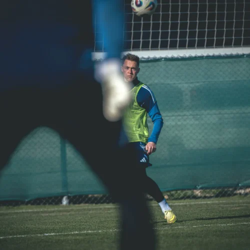 Na zdjęciu widać trzech piłkarzy na boisku, z jednym zawodnikiem w żółtej kamizelce, który obserwuje lecącą piłkę.