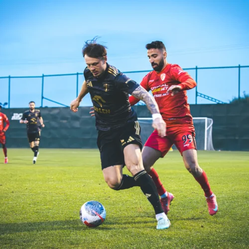 Na zdjęciu widoczna jest scena meczu piłkarskiego, w której zawodnik w czarnej koszulce próbuje uciec przed przeciwnikiem w czerwonym stroju.