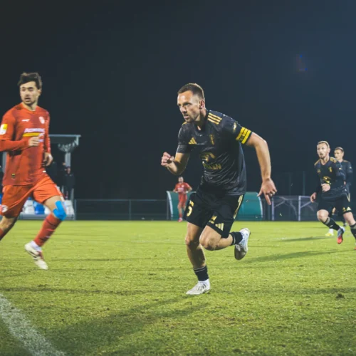 Na zdjęciu widać piłkarzy w trakcie meczu, z wyraźnym skupieniem i dynamiką gry na zielonym boisku w nocy.