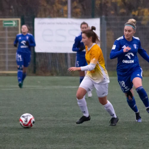 Na zdjęciu widać dwie zawodniczki piłki nożnej biegające po boisku, z piłką znajdującą się blisko jednej z nich.