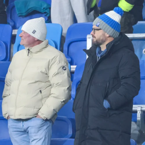 Dwaj mężczyźni w ciepłych kurtkach i czapkach według stoją na trybunach podczas wydarzenia sportowego.
