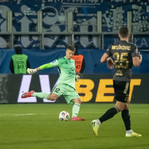 Na zdjęciu bramkarz w zielonym stroju wykonuje wykop piłki na boisku podczas meczu piłkarskiego.